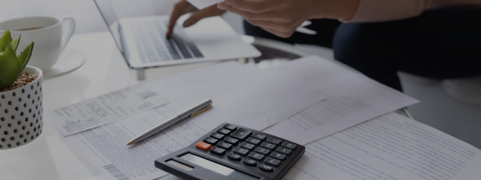 A table with papers strewn across it, a calculator, and a laptop. A person sits behind the table, face out of frame, working. 