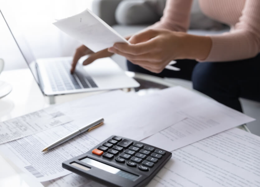 A table with papers strewn across it, a calculator, and a laptop. A person sits behind the table, face out of frame, working. 