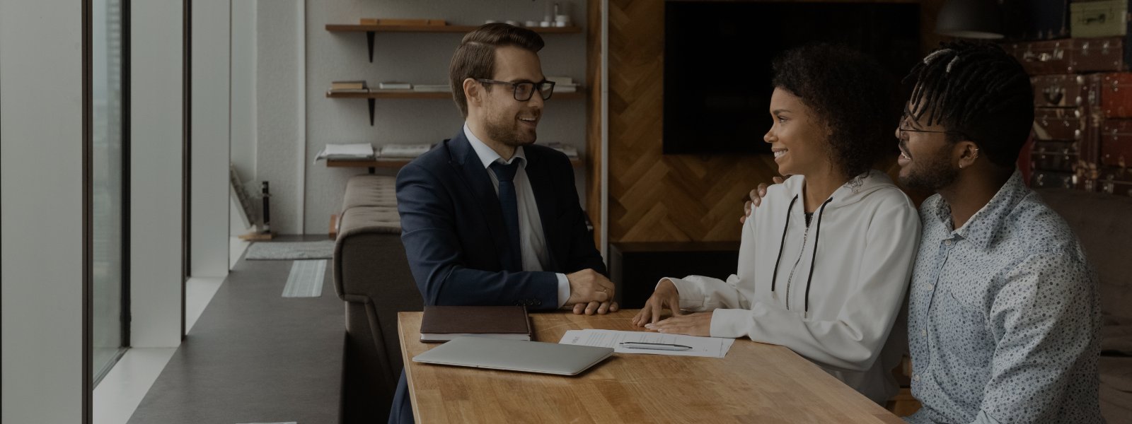 An agent and two clients, seated at a table. The agent and one client are shaking hands. 