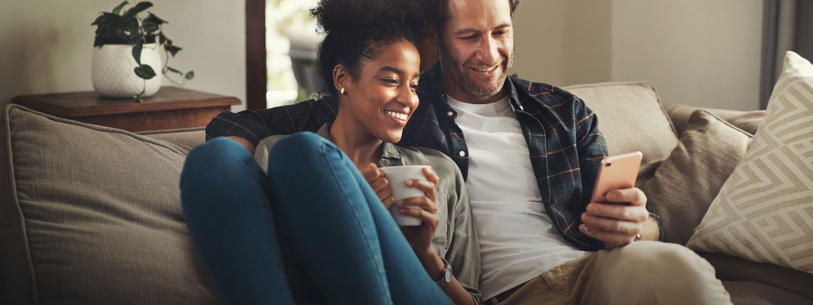 A smiling man and woman sitting on a couch, looking at the phone in the man's hand. 